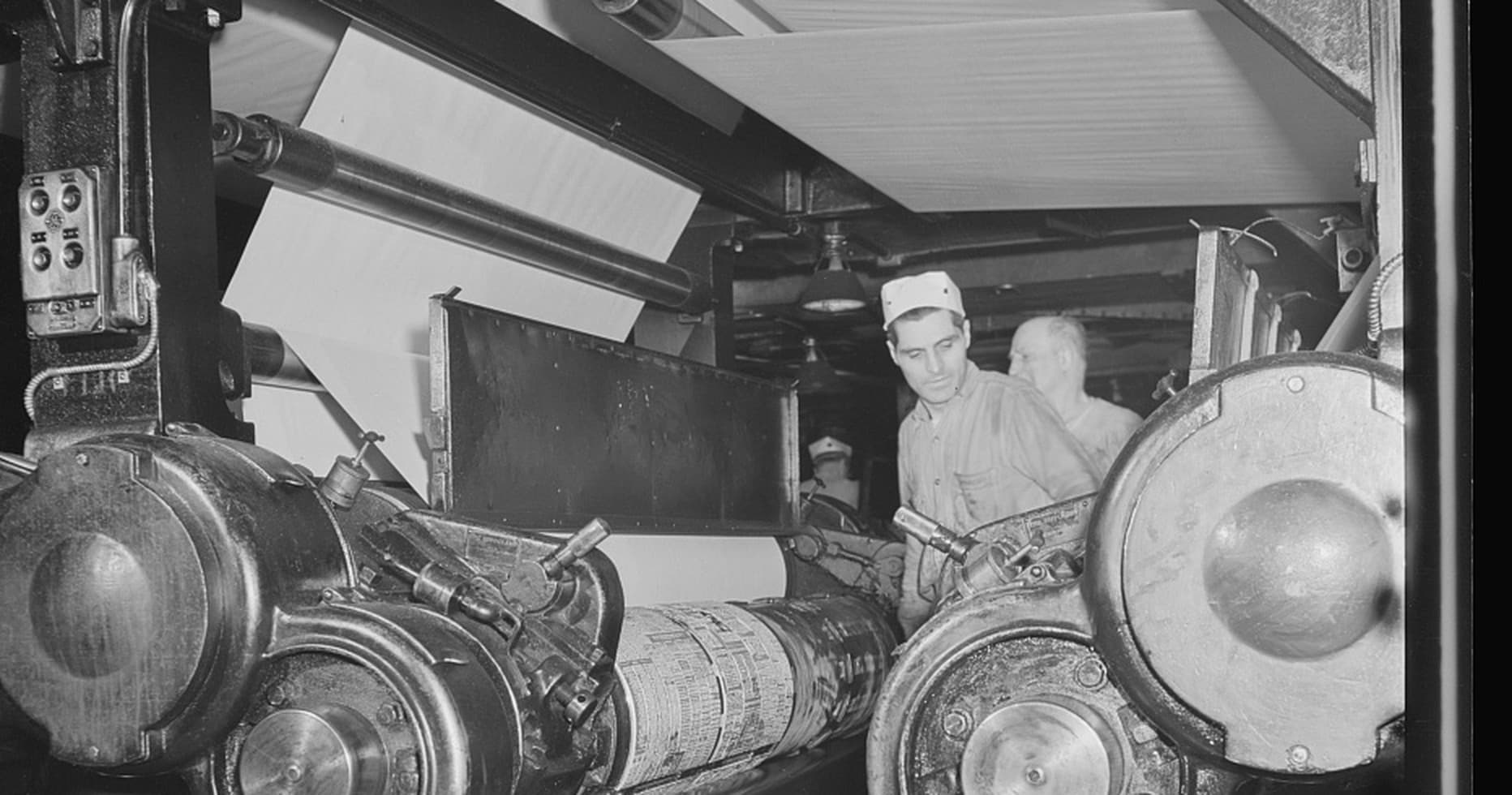 Pressmen running the giant printing presses of a 1942 newspaper pressroom, webs of newsprint overhead